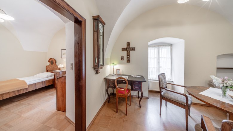 Ein traditionell eingerichtetes Zimmer mit einem Schreibtisch, Stuhl, Tisch und Bett. Ein Kreuz h&auml;ngt an der Wand, und ein Fenster l&auml;sst Tageslicht herein.