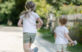 Zwei Kinder laufen auf einem Kiesweg in einem Garten, umgeben von B&auml;umen und Blumen.