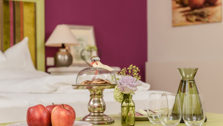 Ein stilvoll eingerichtetes Zimmer mit einem Tisch, auf dem &Auml;pfel, Kekse unter einer Glasglocke, eine Vase mit Blume und Gl&auml;ser stehen.