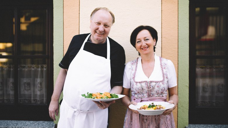 Ein Mann in Schürze und eine Frau in traditioneller Kleidung halten Teller mit Essen vor einem Gebäude.