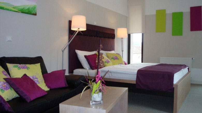 Modern eingerichtetes Hotelzimmer mit Doppelbett, Sofa und Dekoration in Lila- und Gr&uuml;nt&ouml;nen.
