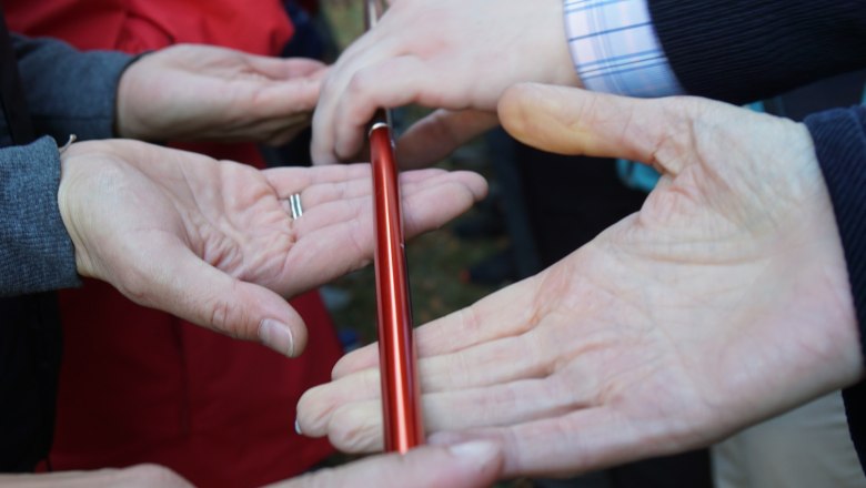 Menschen halten gemeinsam einen roten Stab mit den Fingern.