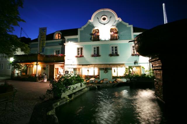 Ein traditionelles Gasthaus mit blauer Fassade und beleuchteten Fenstern bei Nacht, im Vordergrund ein Brunnen.
