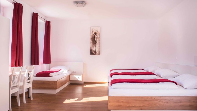 Helles Zimmer mit zwei Betten, roten Vorh&auml;ngen und einem Bild an der Wand.