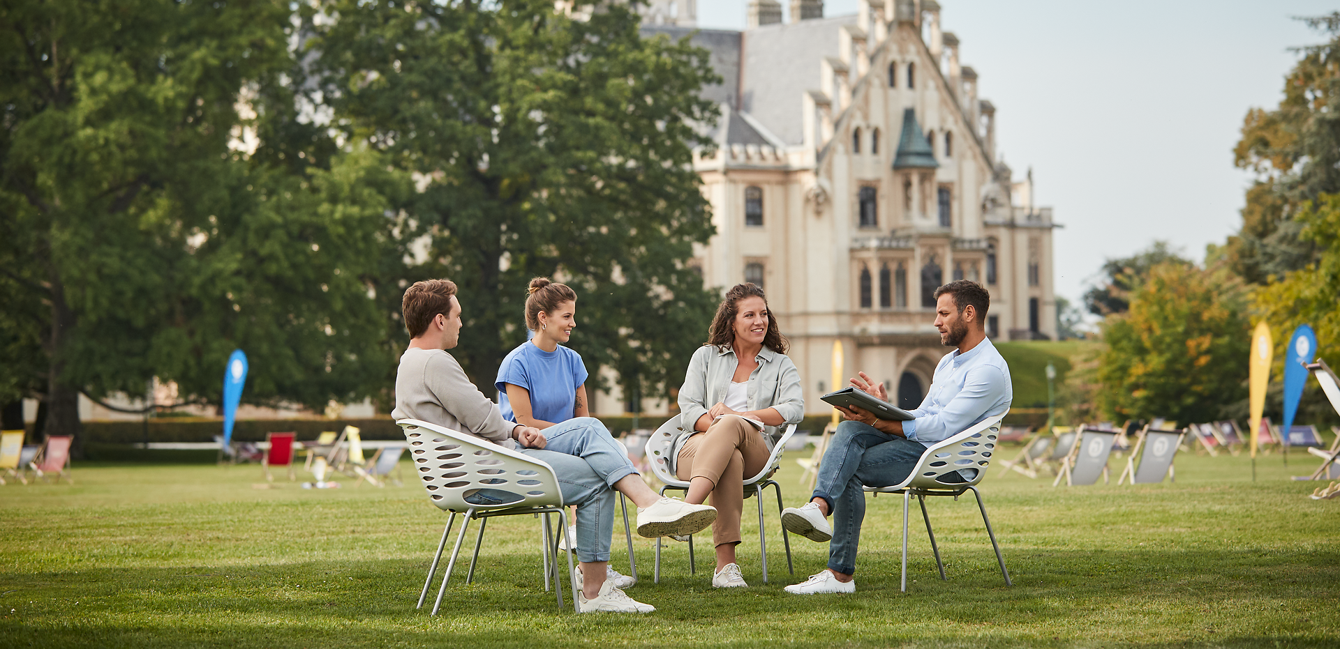 Inmitten einer malerischen Kulisse entspannen sich vier Freunde auf modernen Stühlen, umgeben von üppigem Grün und der majestätischen Architektur im Hintergrund. Die sanfte Brise und das Lachen der Gruppe schaffen eine einladende Atmosphäre, die den perfekten Rahmen für unvergessliche Momente bietet.
