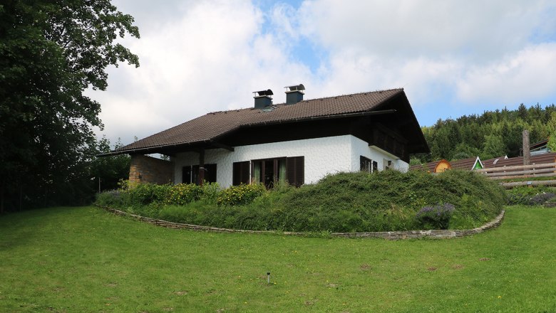 Ein wei&szlig;es Haus mit braunem Dach in einem gr&uuml;nen Garten mit B&auml;umen und Blumen.