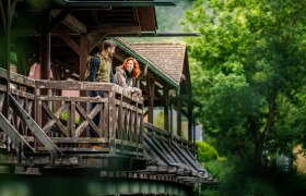Ein Paar steht auf einer &uuml;berdachten Holzbr&uuml;cke und schaut sich an, umgeben von gr&uuml;ner Natur.