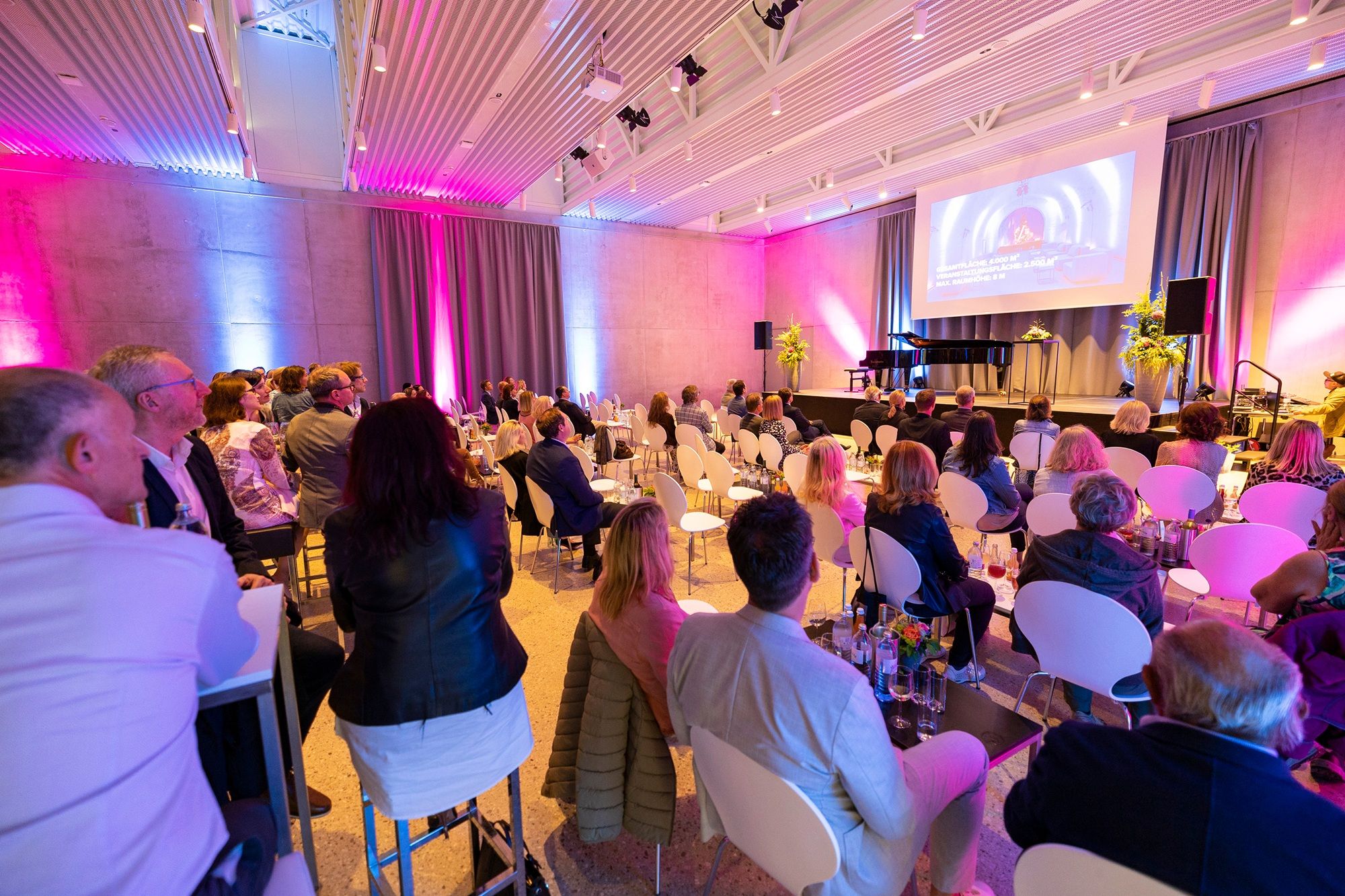 Menschen sitzen in einem modernen Veranstaltungsraum mit Bühne und Leinwand.