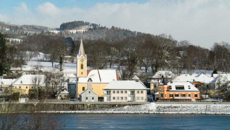 Winterlandschaft mit Kirche und schneebedeckten Häusern am Flussufer.