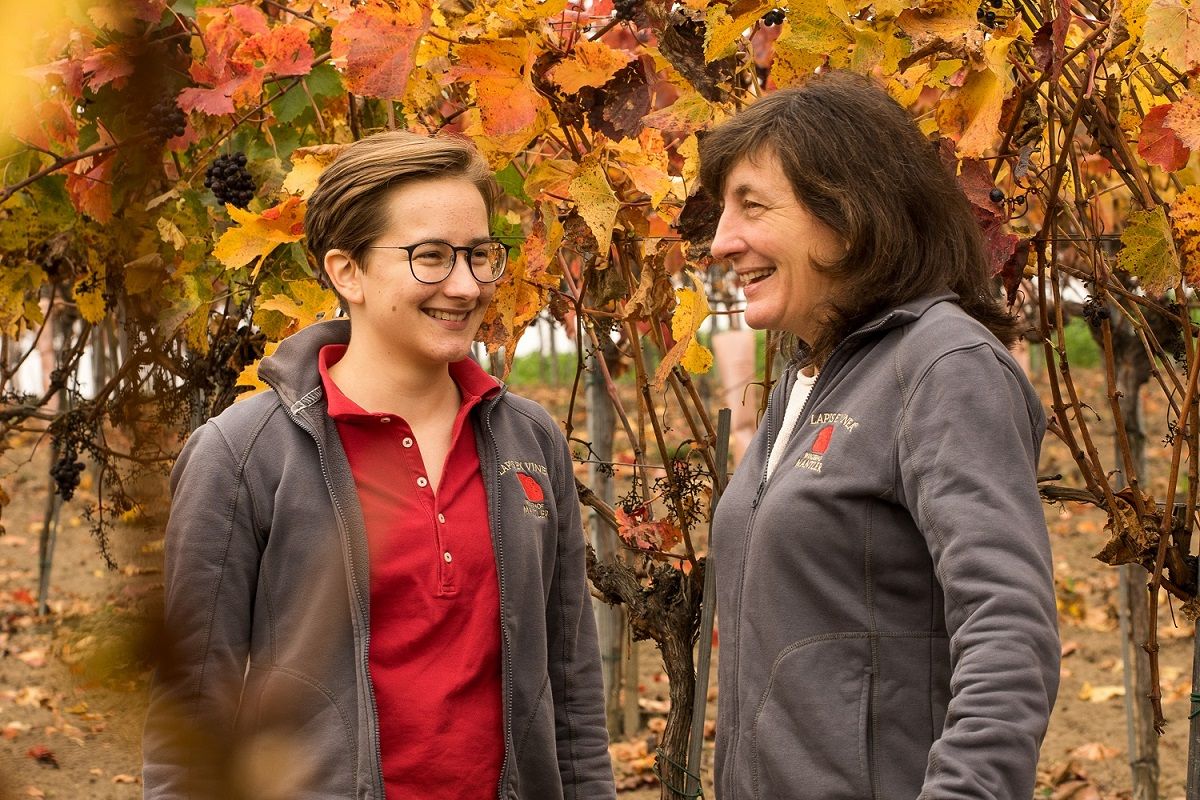 Zwei Frauen stehen lächelnd in einem herbstlichen Weingarten mit bunten Blättern.