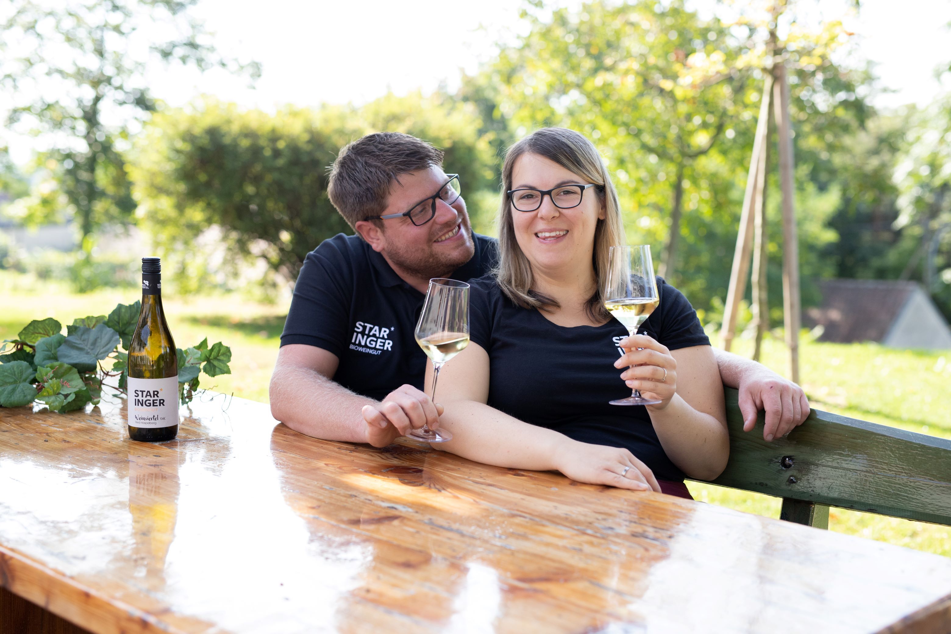 Ein Paar sitzt an einem Holztisch im Freien und hält Weingläser. Eine Weinflasche mit dem Etikett 'Staringer' steht auf dem Tisch.