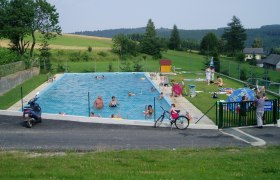Ein Freibad mit mehreren Menschen, umgeben von grüner Landschaft und Bäumen.