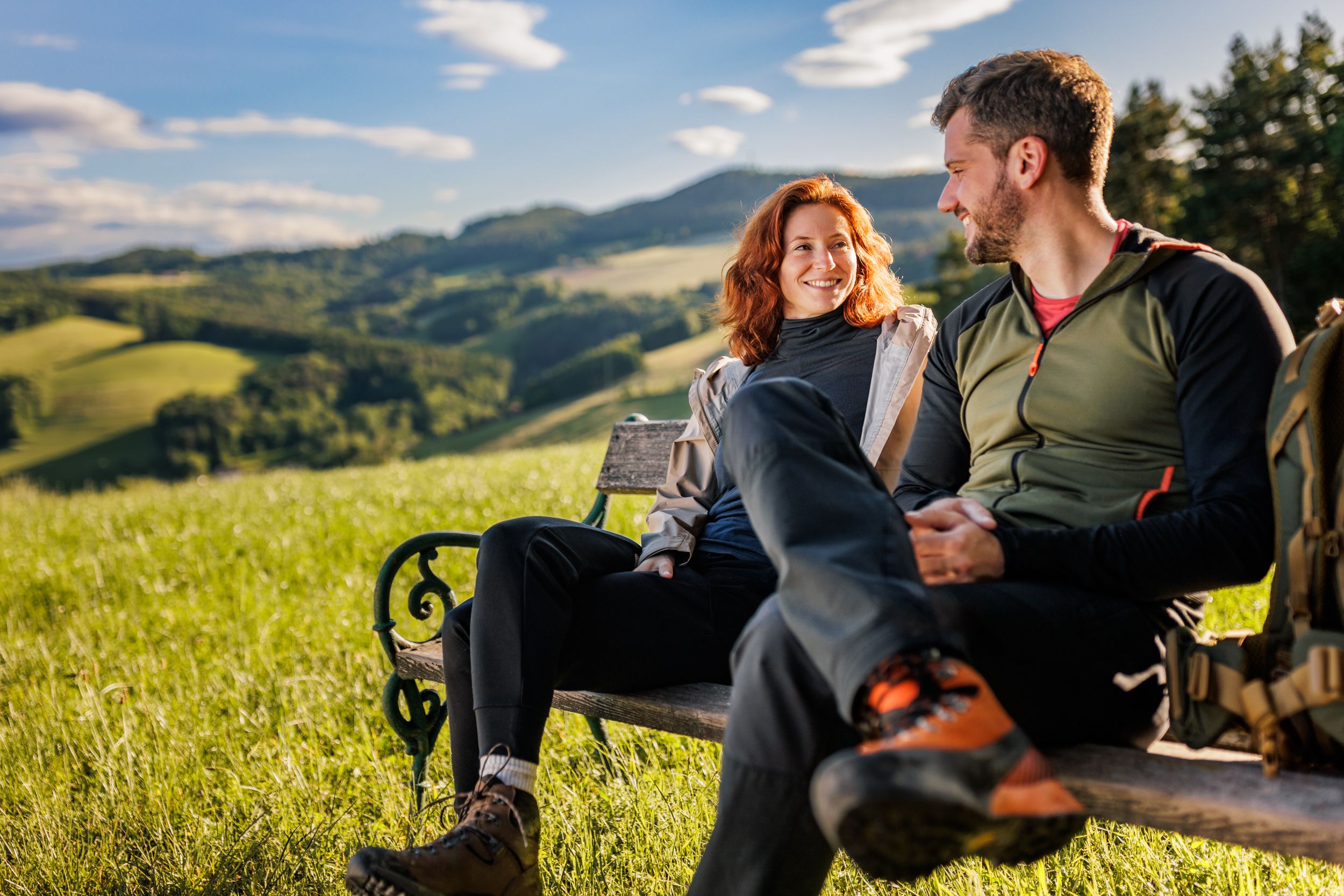 Eine Frau und ein Mann in Wanderkleidung sitzen auf einer Holzbank. Im Hintergrund Wiese, grüne Hügel und Wald.