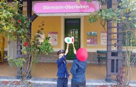 Zwei Kinder in Eisenbahnuniformen stehen vor dem Bahnhof Dürnstein-Oberloiben und halten Signaltafeln hoch.