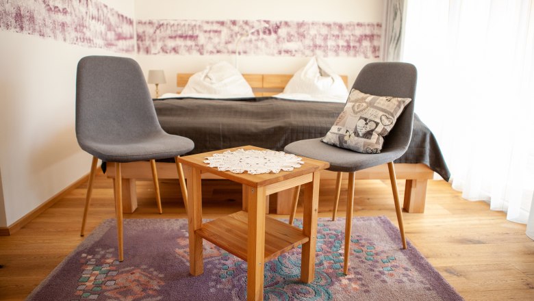 Zwei graue St&uuml;hle und ein Holztisch mit Deckchen in einem Schlafzimmer.