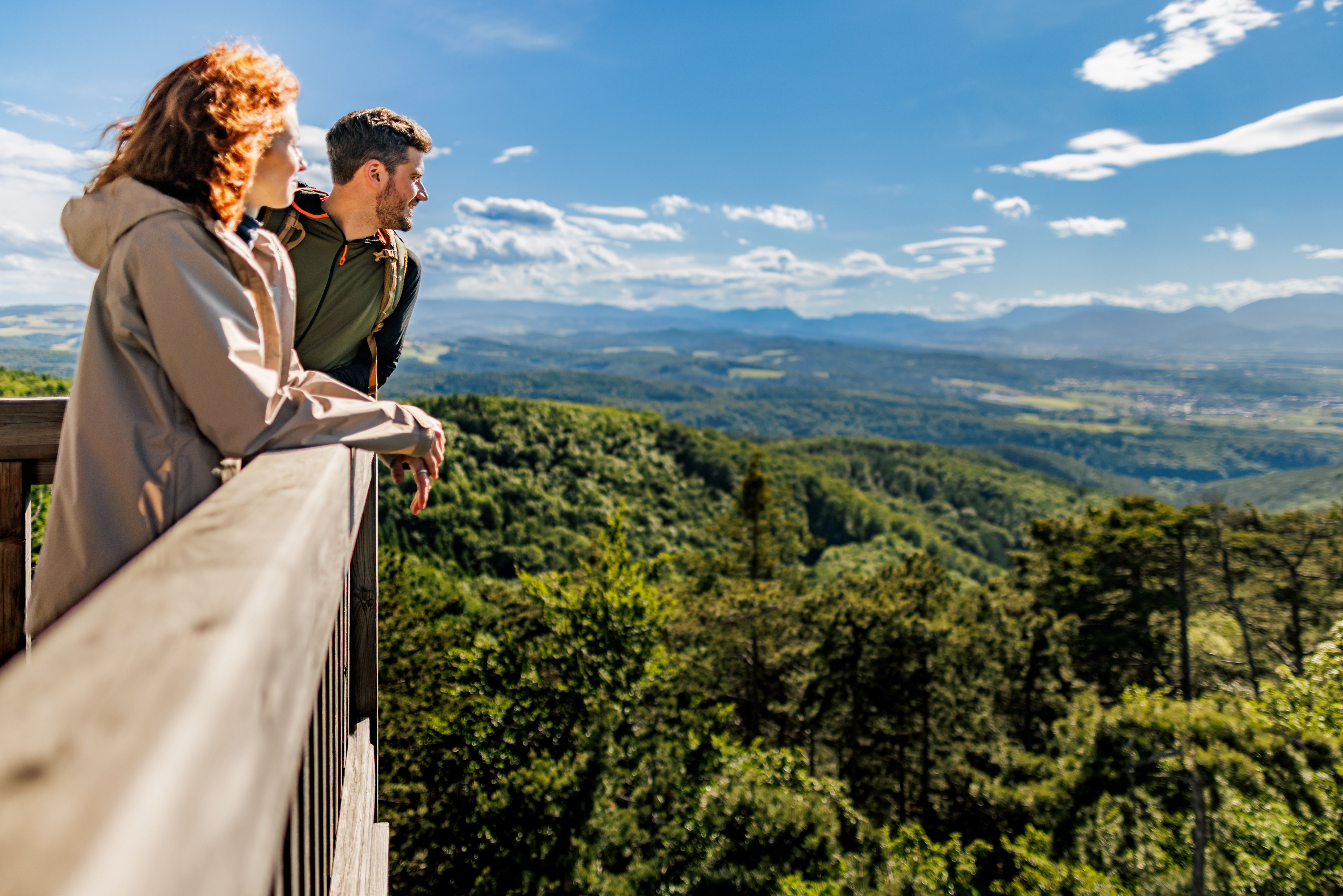 Zwei Personen stehen auf der Aussichtswarte und blicken über eine bewaldete, hügelige Landschaft.