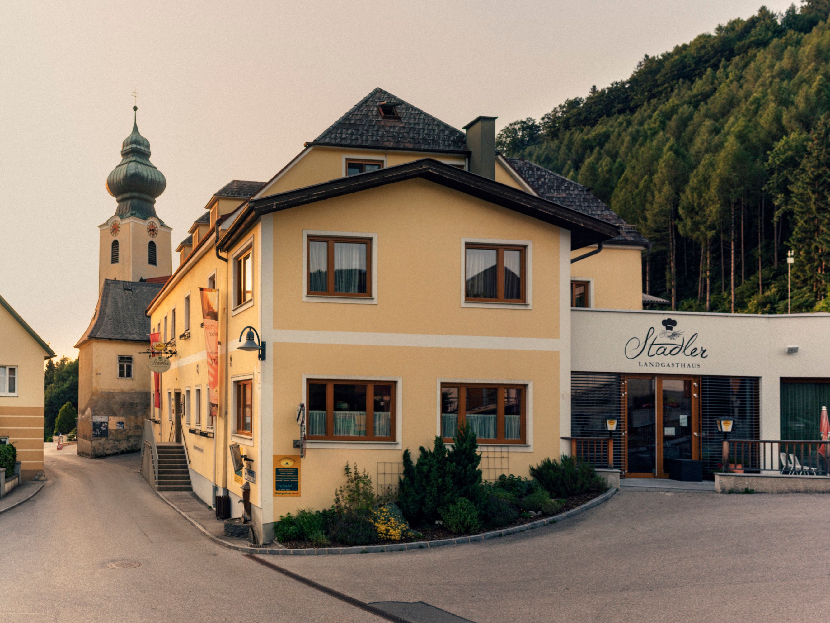 Gelbes Gebäude mit der Aufschrift 'Stadler Landgasthaus' und einer Kirche im Hintergrund.