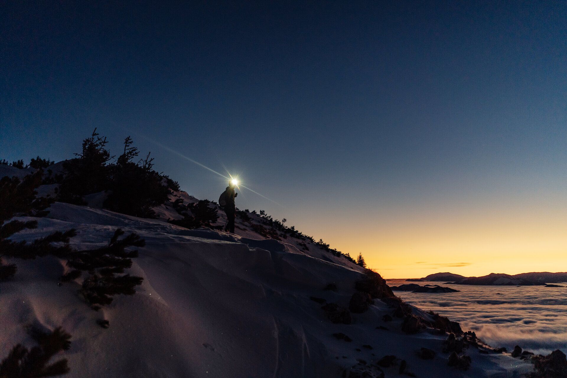 Skitourengeher mit Stirnleuchte vor Sonnenaufgang auf einem verschneiten Berggrat; im Hintergrund weite Winter-Berglandschaft über einem Meer aus Wolken.