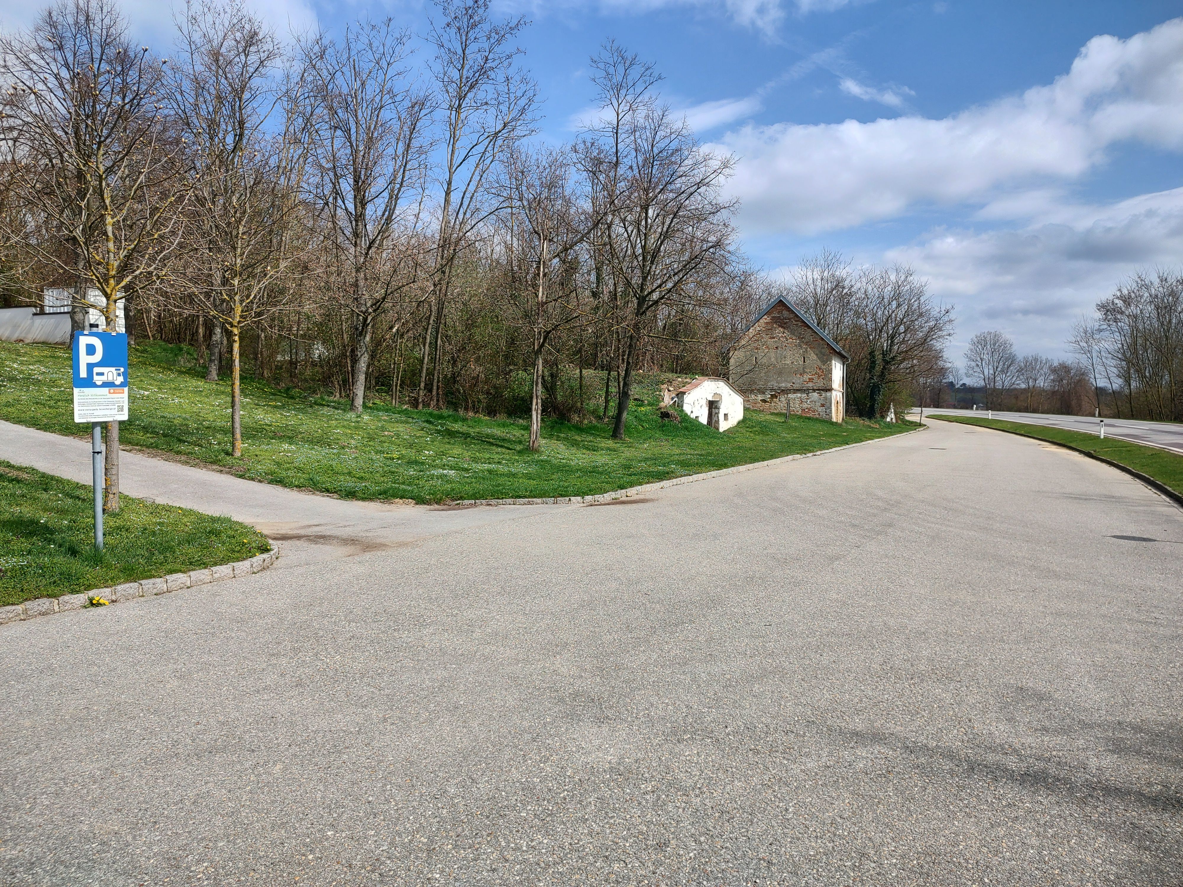 Parkplatz mit Wohnmobilstellplatzschild, Bäume und altes Gebäude im Hintergrund.