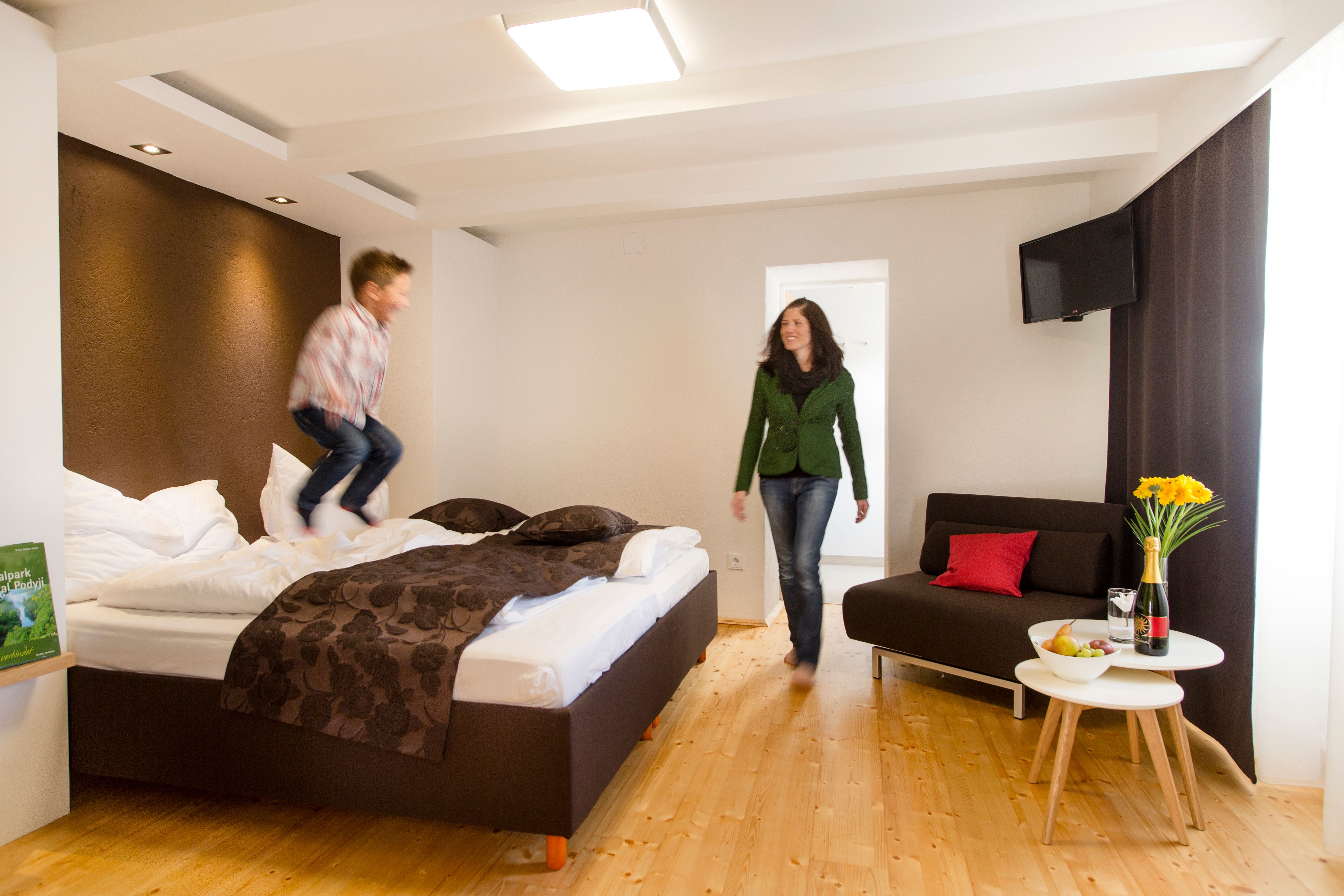 Ein Kind springt auf einem Bett in einem modernen Hotelzimmer, während eine Frau lächelnd zusieht. Ein Tisch mit Blumen und Wein steht daneben.