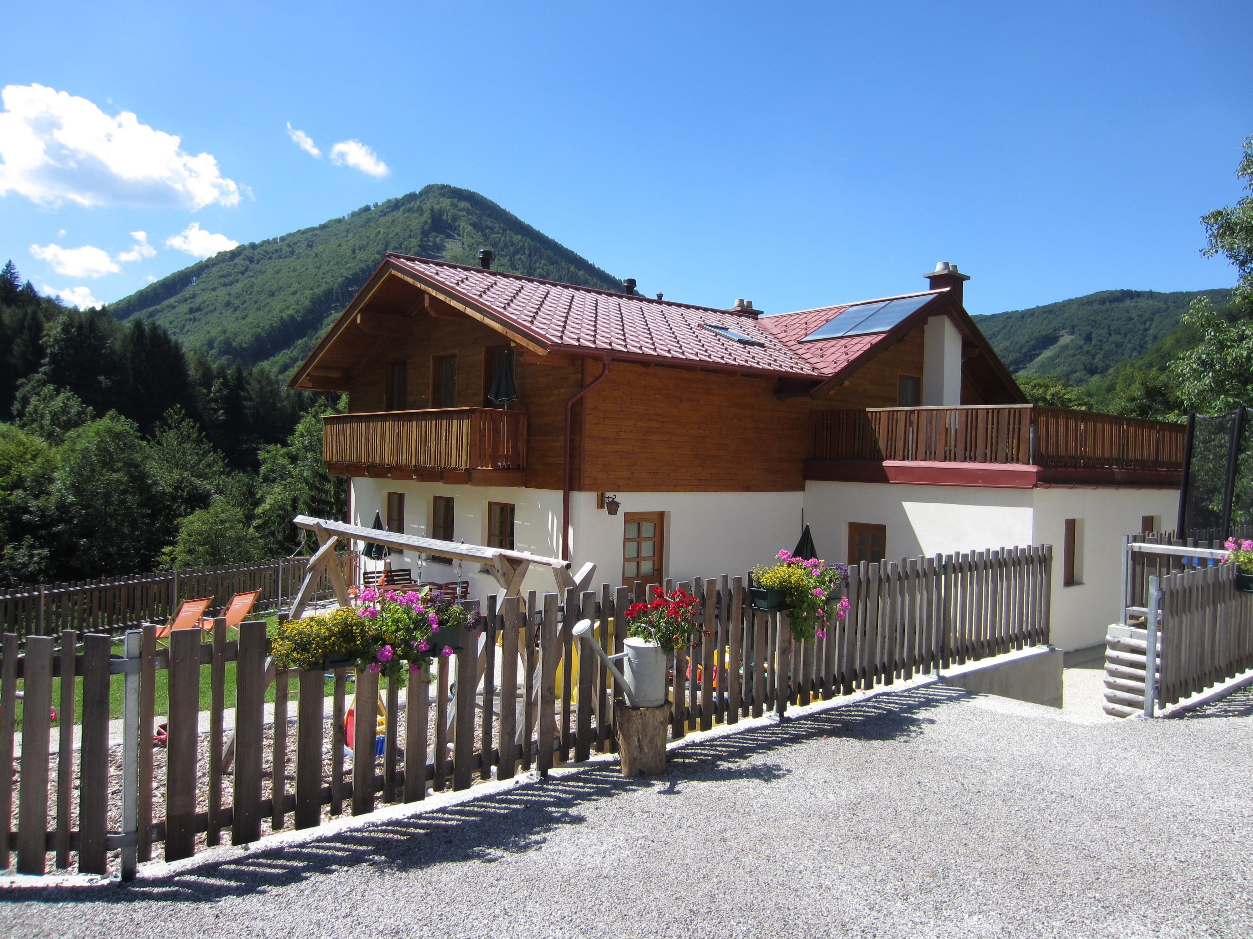 Ein zweistöckiges Haus mit Holzbalkon und Garten, umgeben von Bergen und Bäumen.