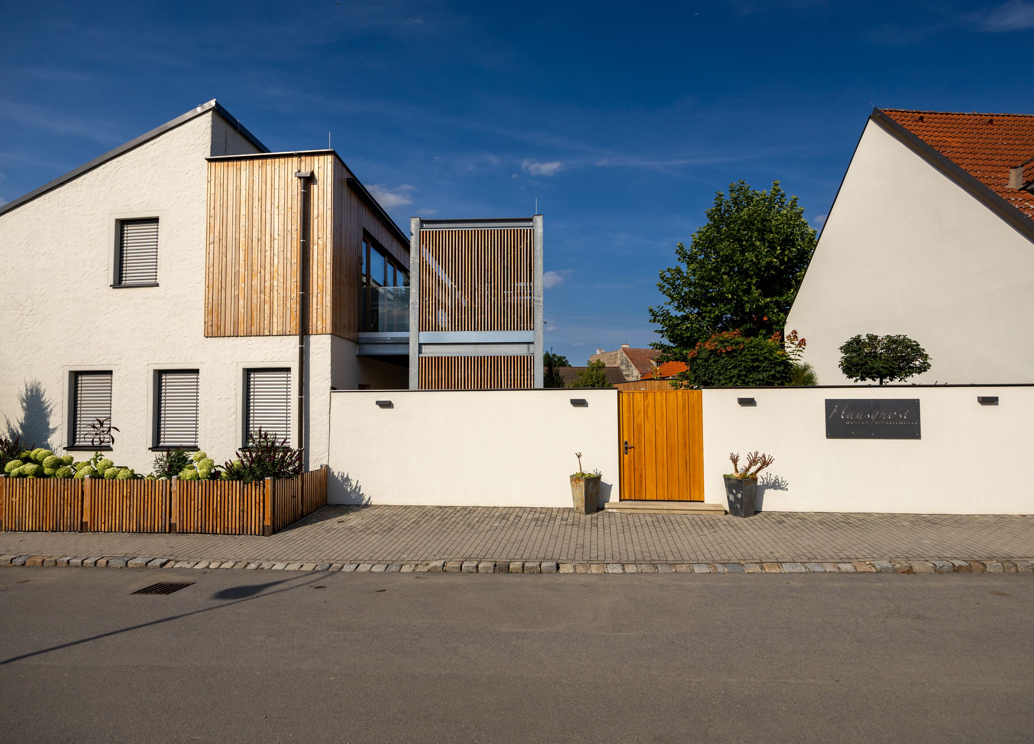 Modernes Gebäude mit weißer Fassade und Holzelementen, umgeben von einer Mauer mit Holztor.