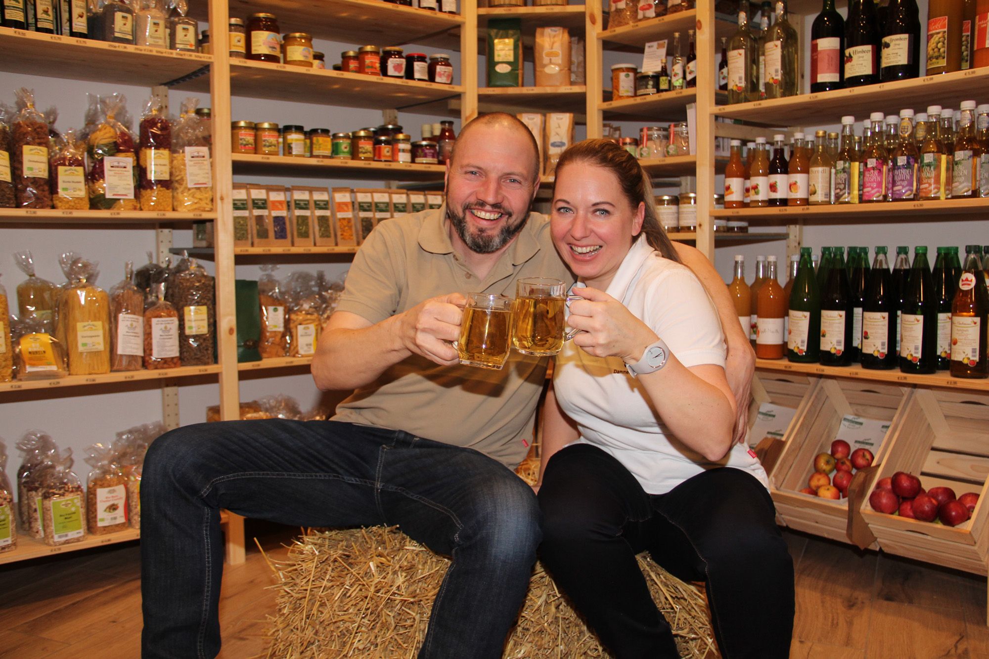 Zwei Personen sitzen auf einem Heuballen im Hofladen und stoßen mit Getränken an.