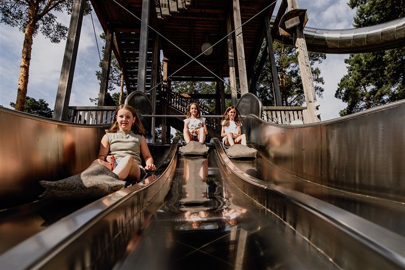 Drei Kinder rutschen auf einer breiten Metallrutsche in einem Erlebnispark.