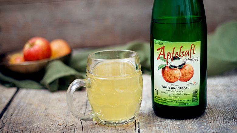 Eine Flasche Apfelsaft und ein Glas gefüllt mit Apfelsaft auf einem Holztisch, im Hintergrund ein grünes Tuch.