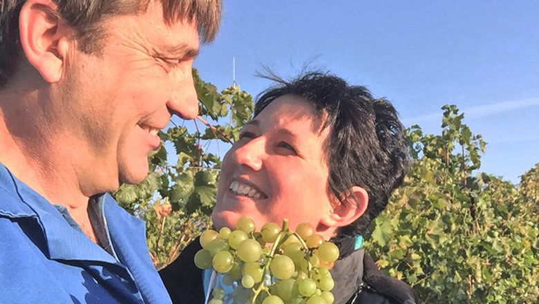 Ein Mann und eine Frau lächeln sich in einem Weinberg an, während sie eine Traube halten.