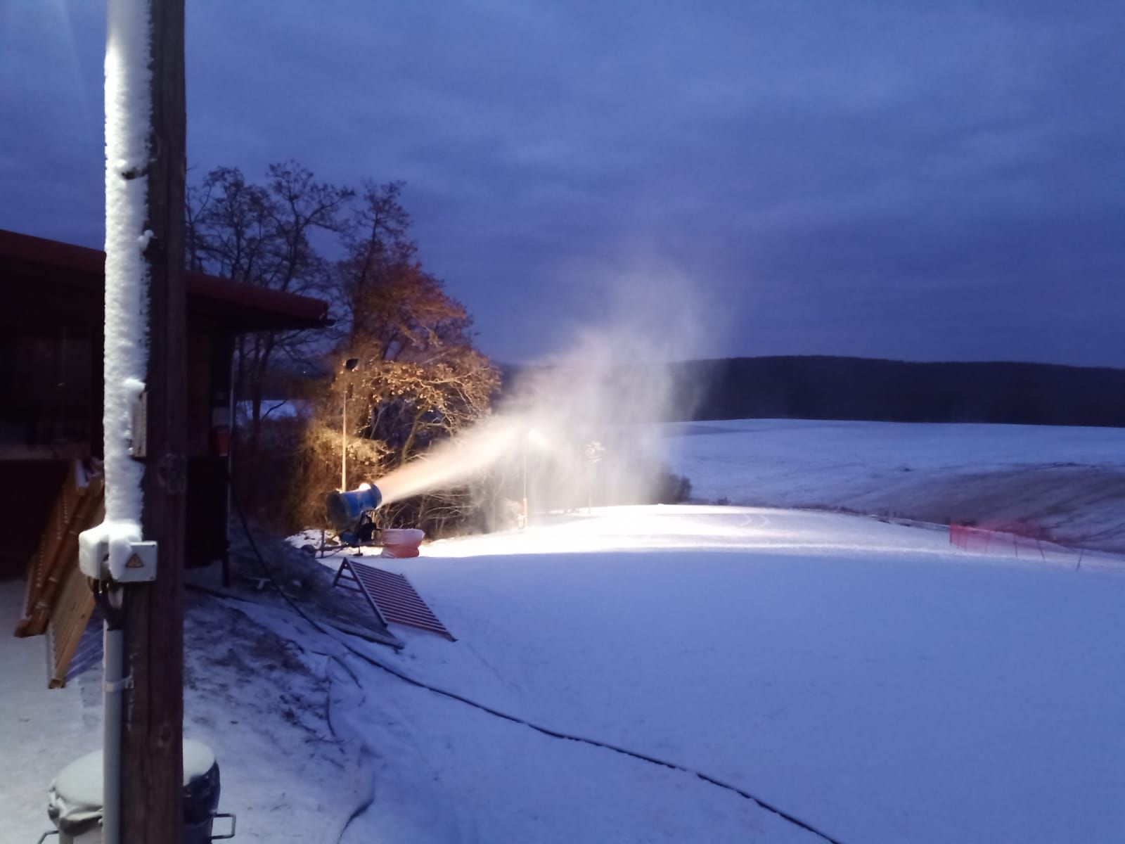 Schneekanone beschneit eine Piste bei Dämmerung.