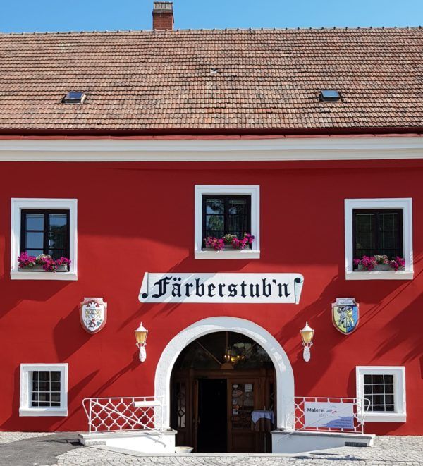 Rotes Gebäude mit der Aufschrift 'Färberstub'n' und blühenden Blumen in den Fenstern.