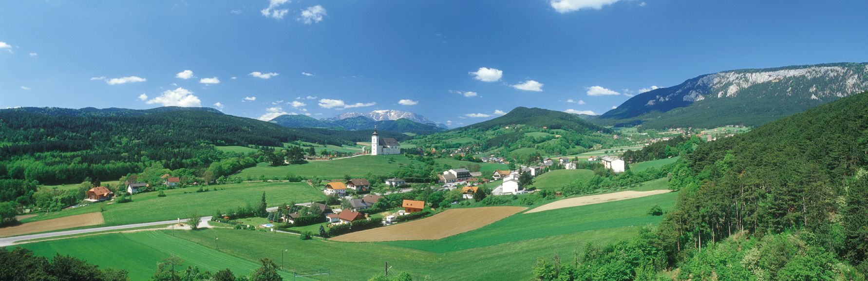 Panoramablick auf Höflein mit Kirche, grünen Feldern und Bergen im Hintergrund.