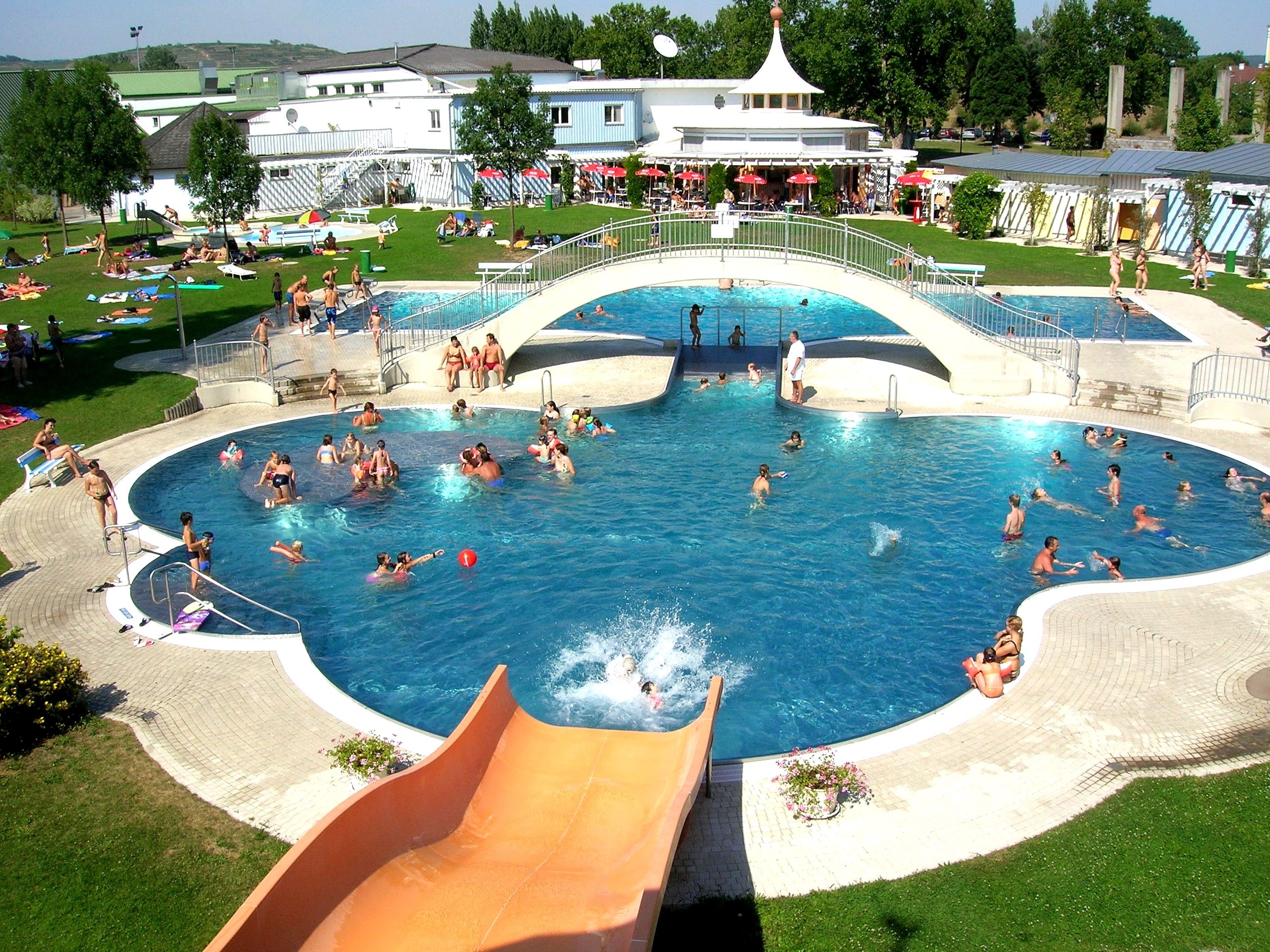 Freibad mit Pool, Brücke und Rutsche, viele Menschen beim Schwimmen und Sonnenbaden.