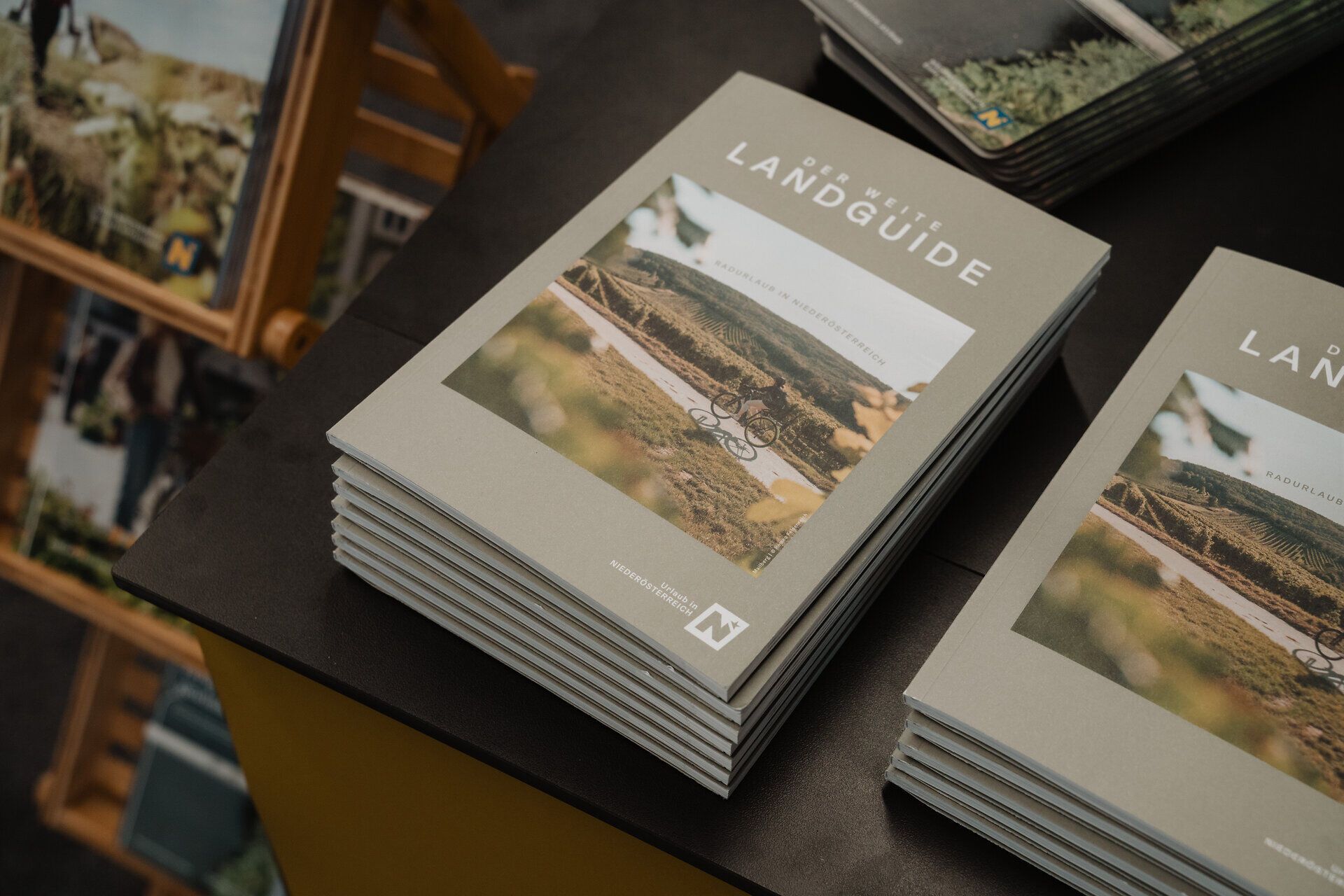 Mehrere Exemplare des Radguides „Der Weite Landguide“ liegen auf einem Tisch. Auf dem Cover ist eine Radfahrerin in einer vom Weinanbau geprägten Landschaft abgebildet.