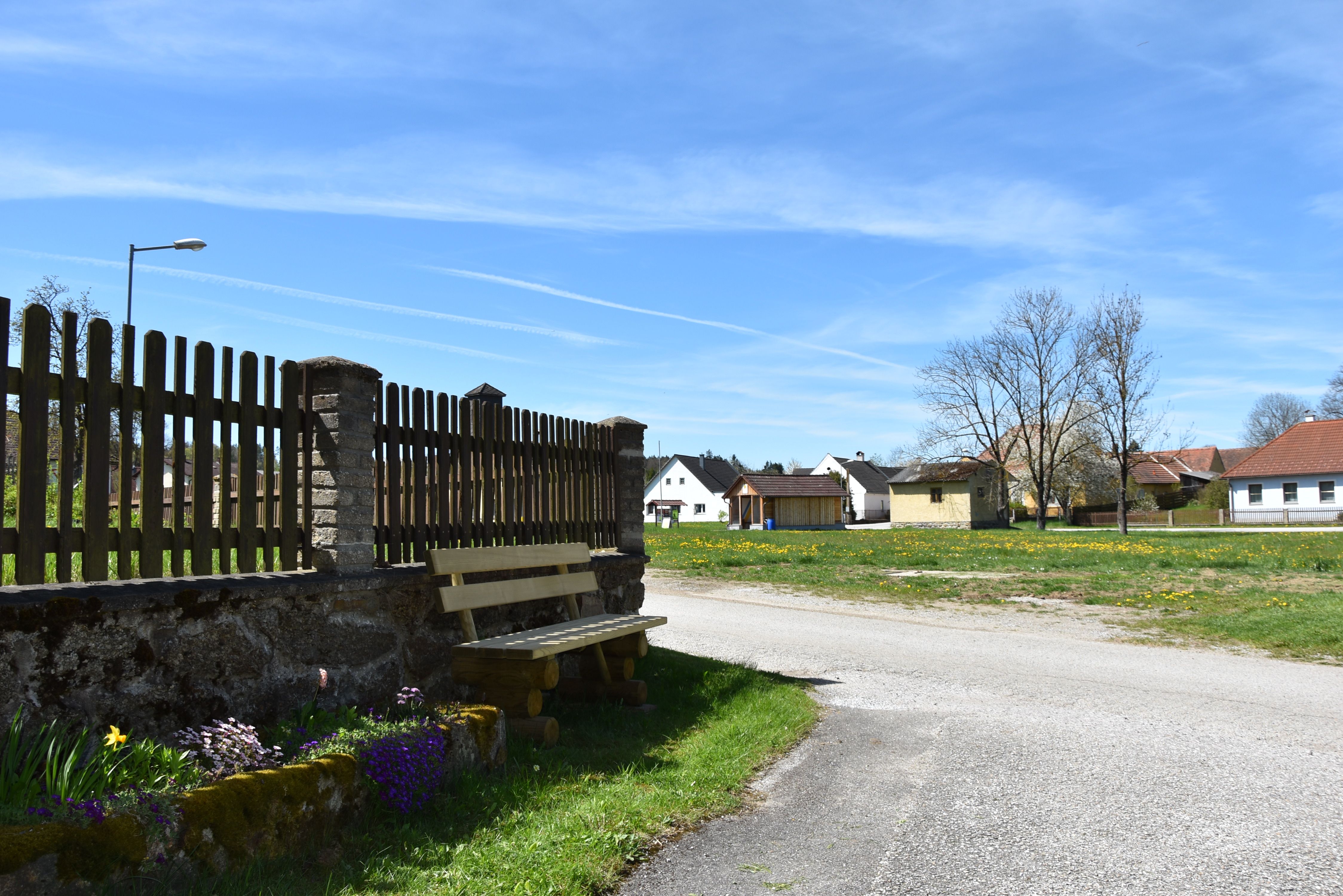Sitzgelegenheit vor dem Haus an einer Dorfstraße mit Zaun, Blumenbeet und Blick auf Häuser unter blauem Himmel.