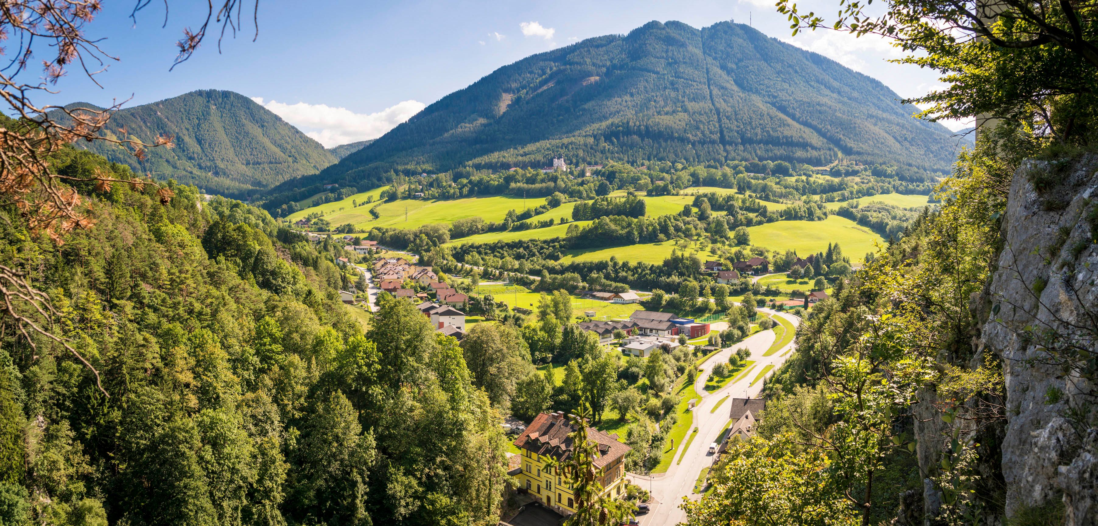 Panoramablick auf ein grünes Tal mit Bergen im Hintergrund und einem Dorf im Vordergrund.
