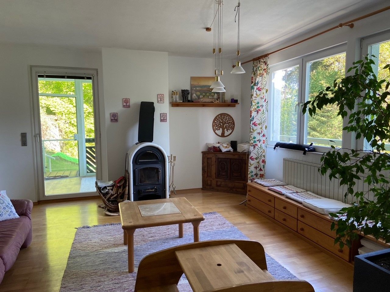 Ein gemütliches Wohnzimmer mit Holzofen, Holzmöbeln und großen Fenstern mit Blick ins Grüne.