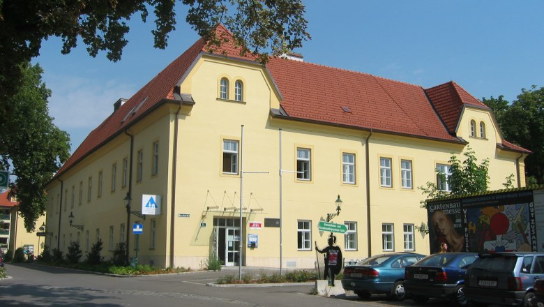 Au&szlig;enansicht eines gelben Geb&auml;udes mit rotem Dach, Junges Hotel Tulln, umgeben von B&auml;umen und parkenden Autos.