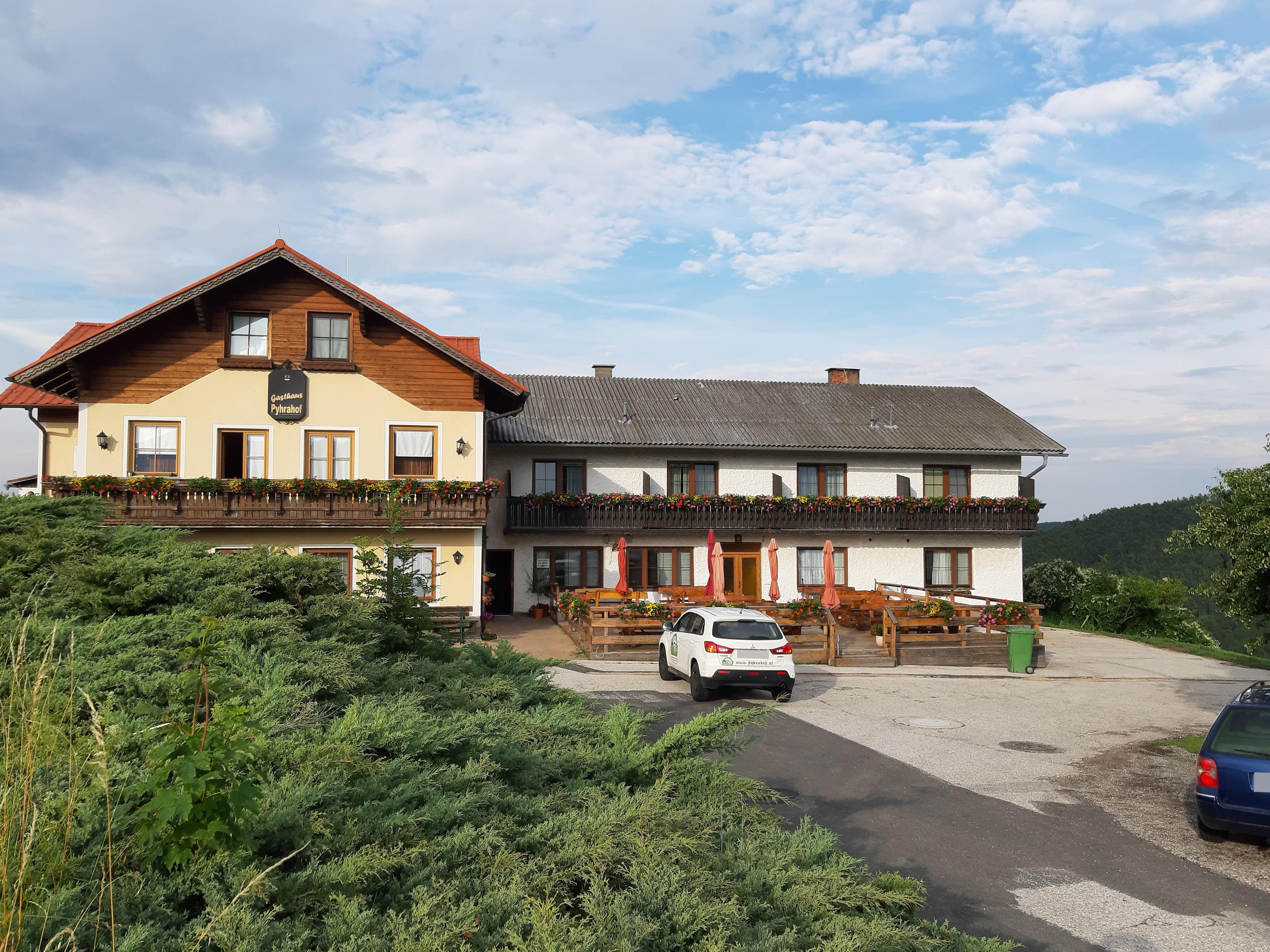 Ein traditionelles Gasthaus mit Balkon und Blumenkästen, davor ein Parkplatz mit Autos, umgeben von grüner Landschaft und blauem Himmel.