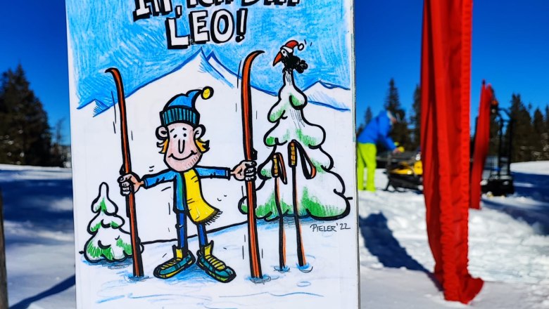 Ein Schild im Schnee mit einer Zeichnung von Leo, einem Cartoon-Charakter mit Skiern, vor einem blauen Himmel.