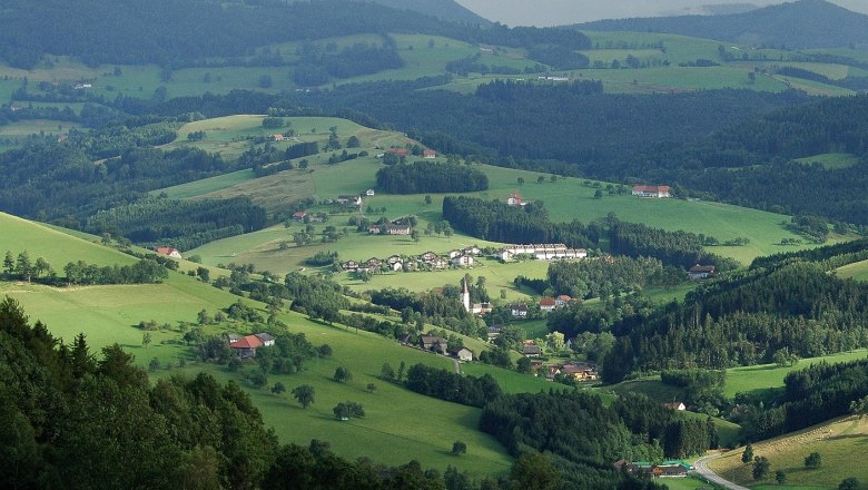 Panoramablick auf eine gr&uuml;ne, h&uuml;gelige Landschaft mit verstreuten H&auml;usern und W&auml;ldern.