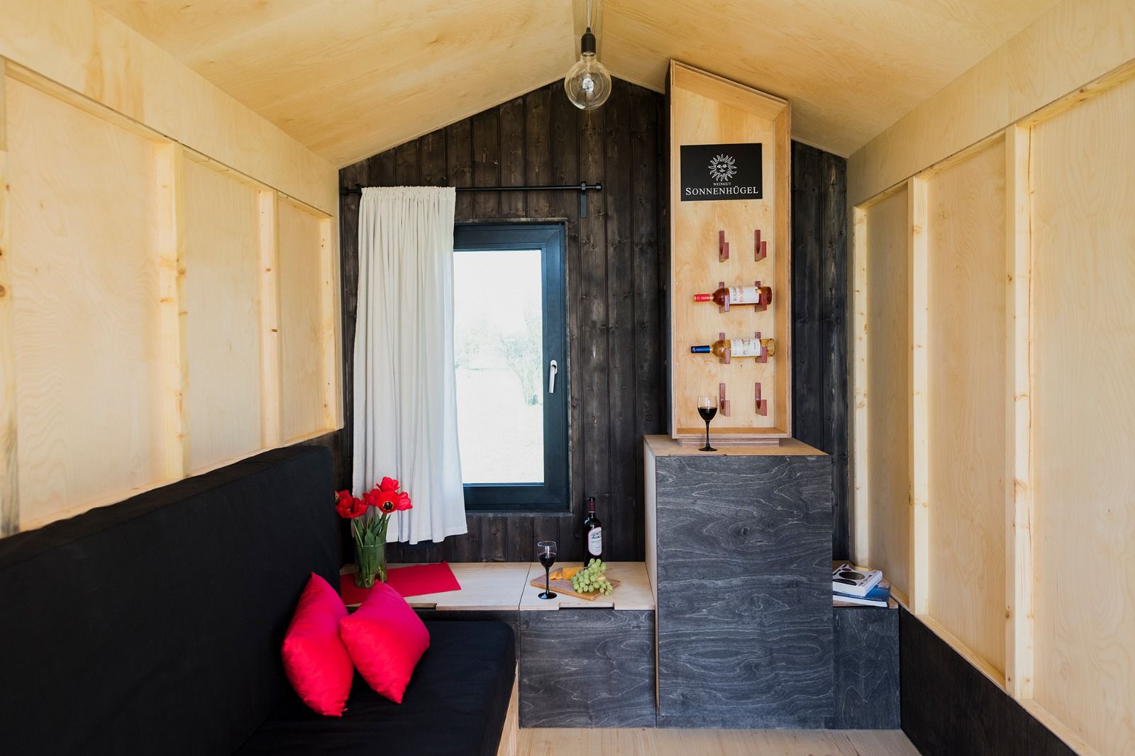 Innenansicht einer gemütlichen Weinberghütte mit Holzwänden, einem Sofa, roten Kissen, einem Weinglas und Weinflaschen.