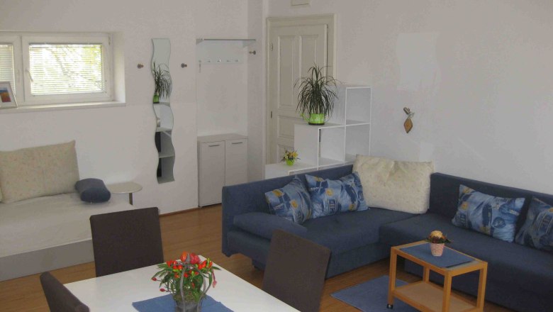 Apartment mit blauen Sofa, weisem Tisch und blauen St&uuml;hlen. Im Hintergrund ein kleines Fenster, T&uuml;r und Spiegel.