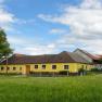 Gelbes Bauernhaus auf einer gr&uuml;nen Wiese mit blauem Himmel und Wolken.