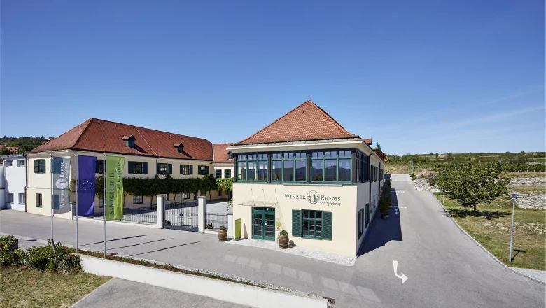 Gebäude der Winzer Krems mit rotem Dach und grünen Fensterläden, umgeben von Weinbergen.