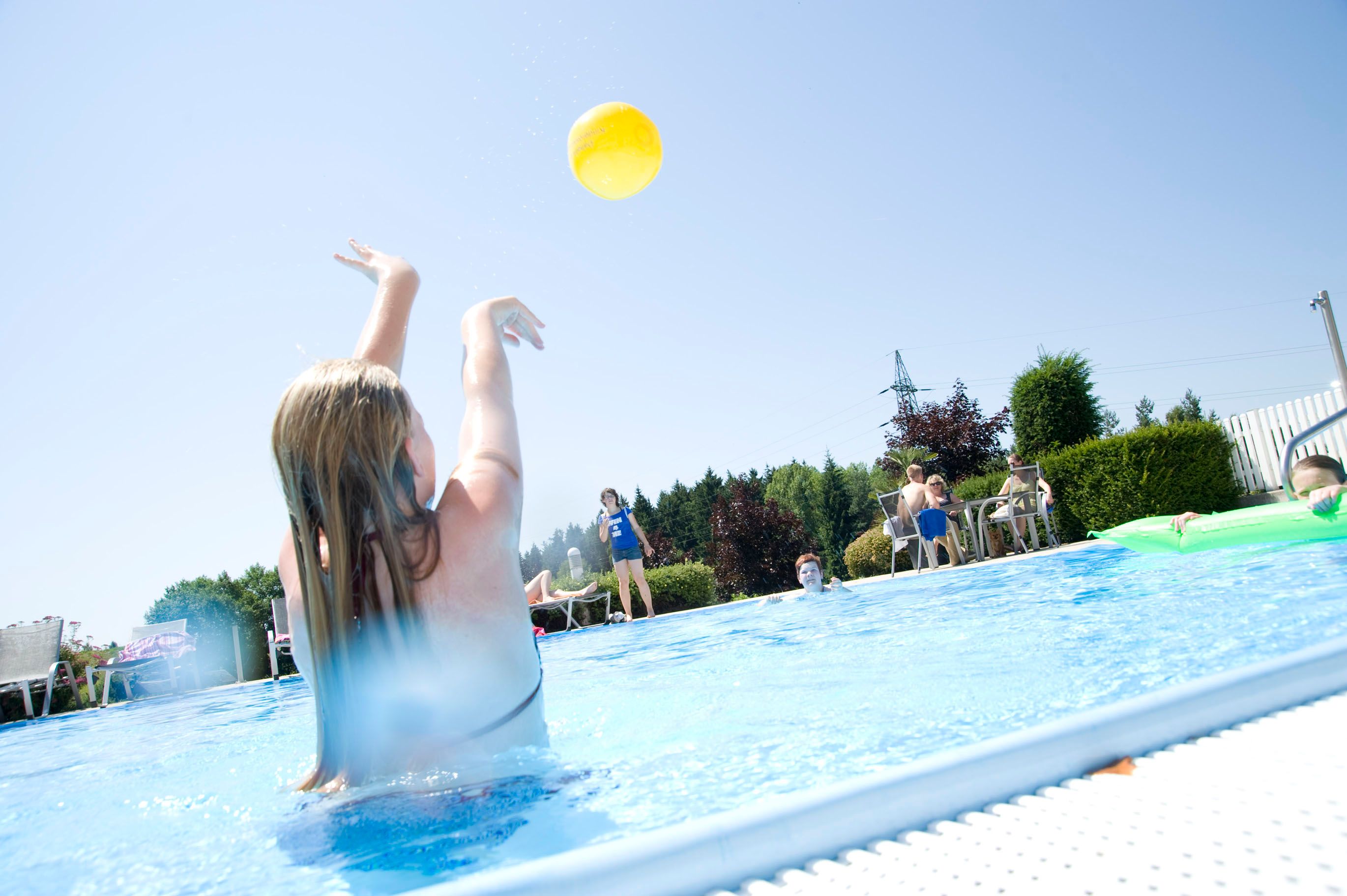 Person im Pool wirft gelben Ball, umgeben von anderen Menschen und Bäumen.