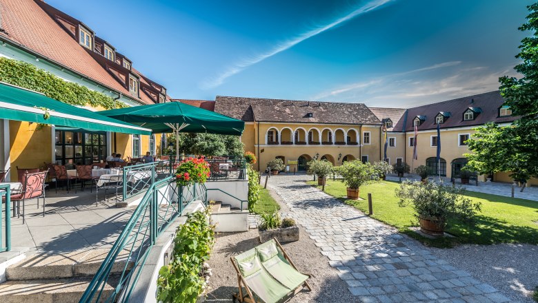 Ein sonniger Innenhof mit gelben Geb&auml;uden, Terrassenbereich und Liegest&uuml;hlen.