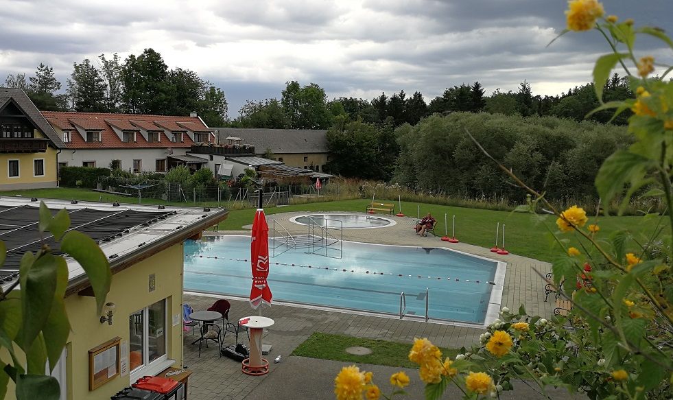 Ein Freibad mit Schwimmbecken, umgeben von Gebäuden und Bäumen, unter bewölktem Himmel.