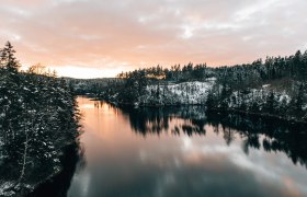 Die sanften Wellen des Stausees spiegeln die schneebedeckten Ufer wider, w&auml;hrend die untergehende Sonne den Himmel in warmen Farben erleuchtet. Eine friedliche Stille umgibt die Landschaft, die zum Verweilen und Entspannen einl&auml;dt.